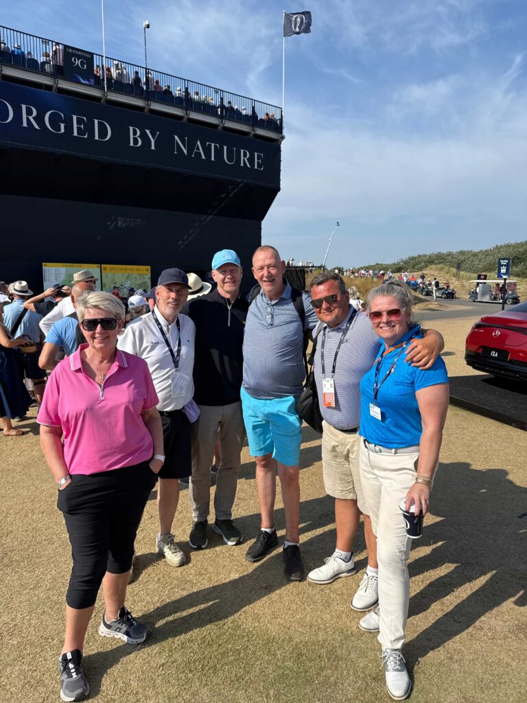 IMG_5278 - Golfsamband Íslands Group of six adults posing together in front of a large dark ship with'FORGED BY NATURE' on its side, at an outdoor event on a sunny day.