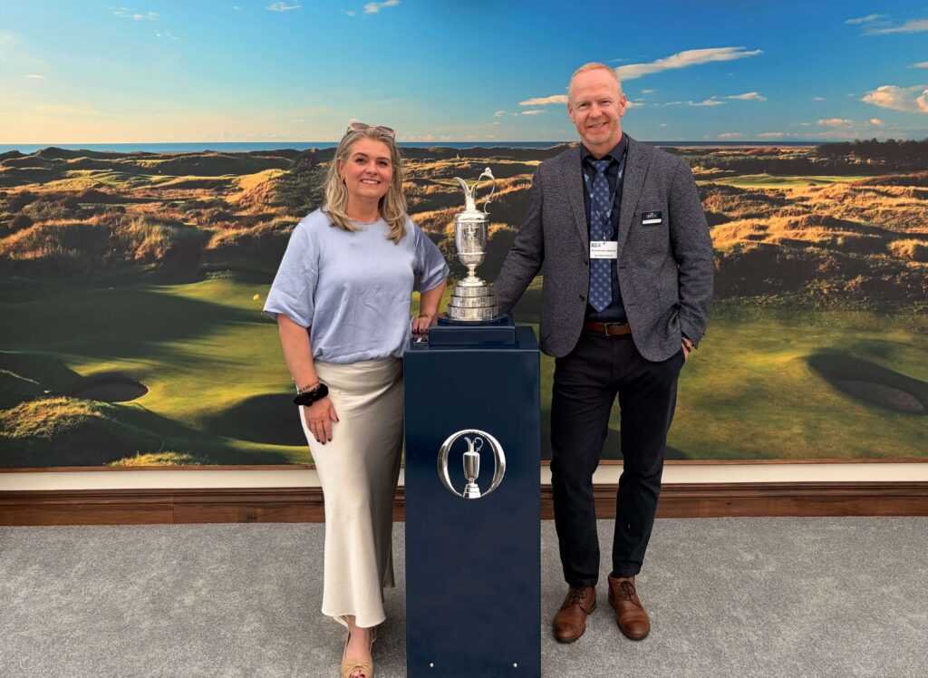 IMG_8381 - Golfsamband Íslands Two people stand beside a tall silver golf trophy, posing in front of a golf course backdrop with blue sky.