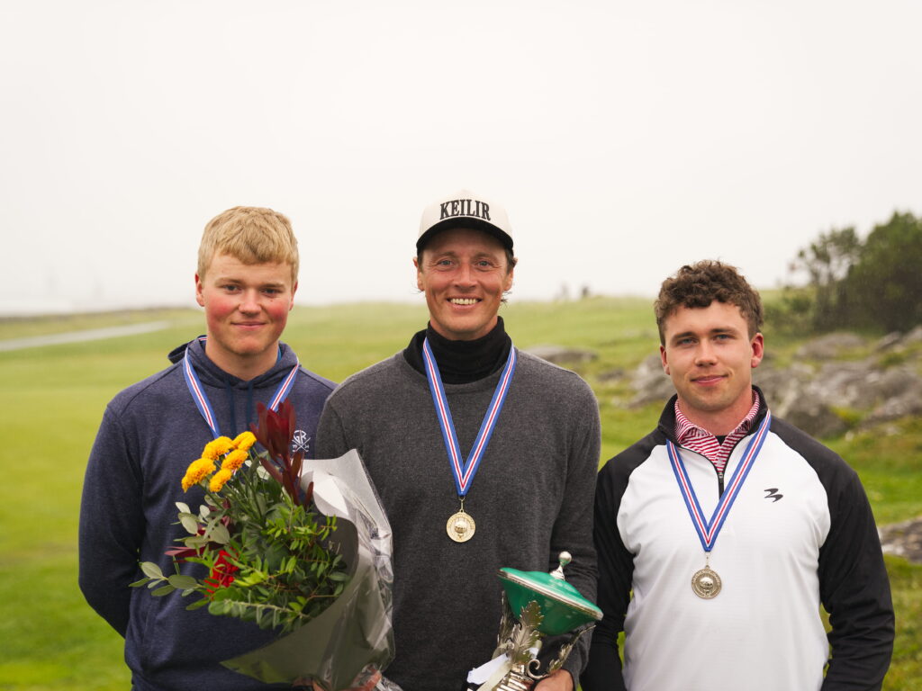 Karlar H - Golfsamband Íslands Three young men posing outdoors with medals around their necks; the center man holds a bouquet of flowers and a trophy, while the others stand beside him on a grassy field.