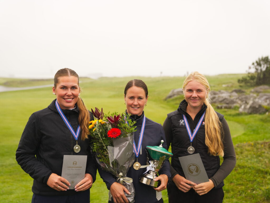 Kvenna H - Golfsamband Íslands Three female athletes stand together outdoors, wearing medals and holding flowers, certificates, and a trophy after a competition.
