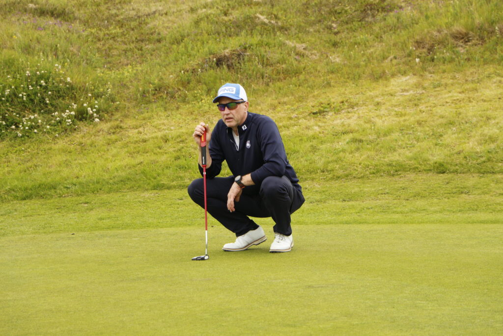 _MG_0913 - Golfsamband Íslands A man in a navy jacket and white cap crouches on a golf green, holding a putter and looking toward the hole with sunglasses on.