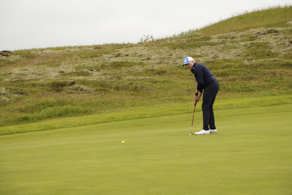 _MG_0928 - Golfsamband Íslands A golfer wearing a white cap and dark outfit lines up a putt on a green, with a grassy hill behind him and a yellow ball near the hole.