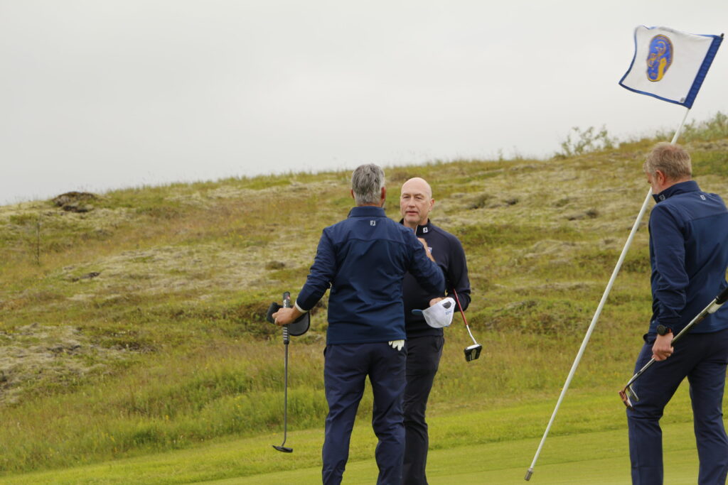 _MG_0938 - Golfsamband Íslands Three men in navy golf outfits on a grassy hill, a blue‑and‑white flag fluttering on a pole nearby.