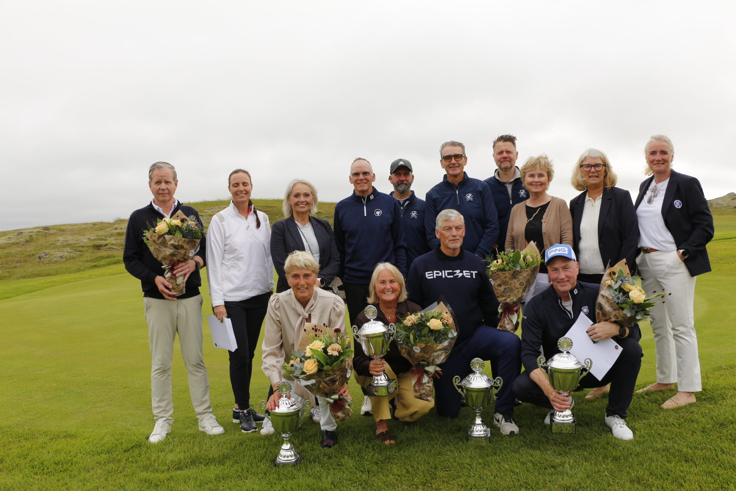 Group of men and women posing on a golf course with large trophies and bouquets after a tournament, cloudy sky overhead.