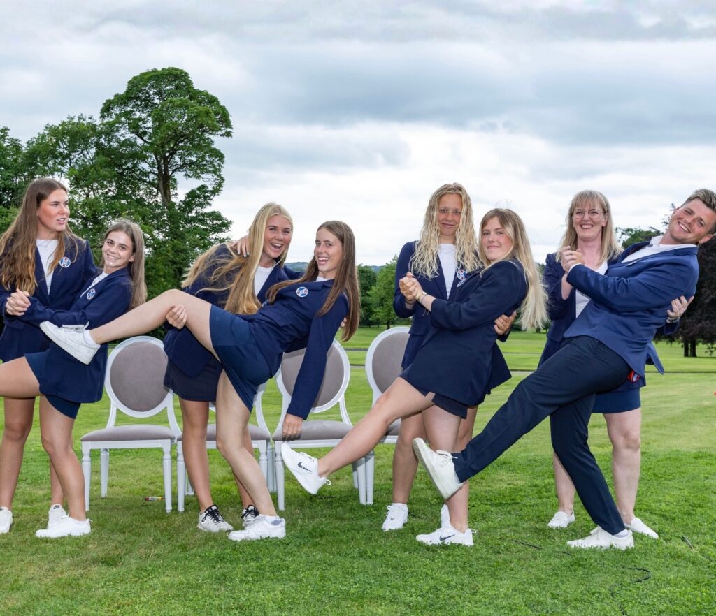 SnapInstto_741454166_18469551109110233_4212386010127251195_n - Golfsamband Íslands Group of students in navy blazers posing outdoors, lifting their legs while standing behind chairs on a grassy field, smiling.
