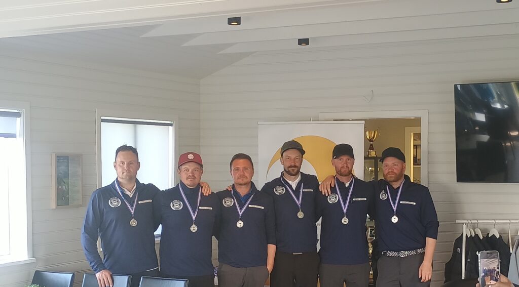 027ca1d5-1dec-40bd-aa97-f876f26ed59b~1 - Golfsamband Íslands Six men in navy uniforms with medals pose side by side indoors, with a banner and trophy in the background.