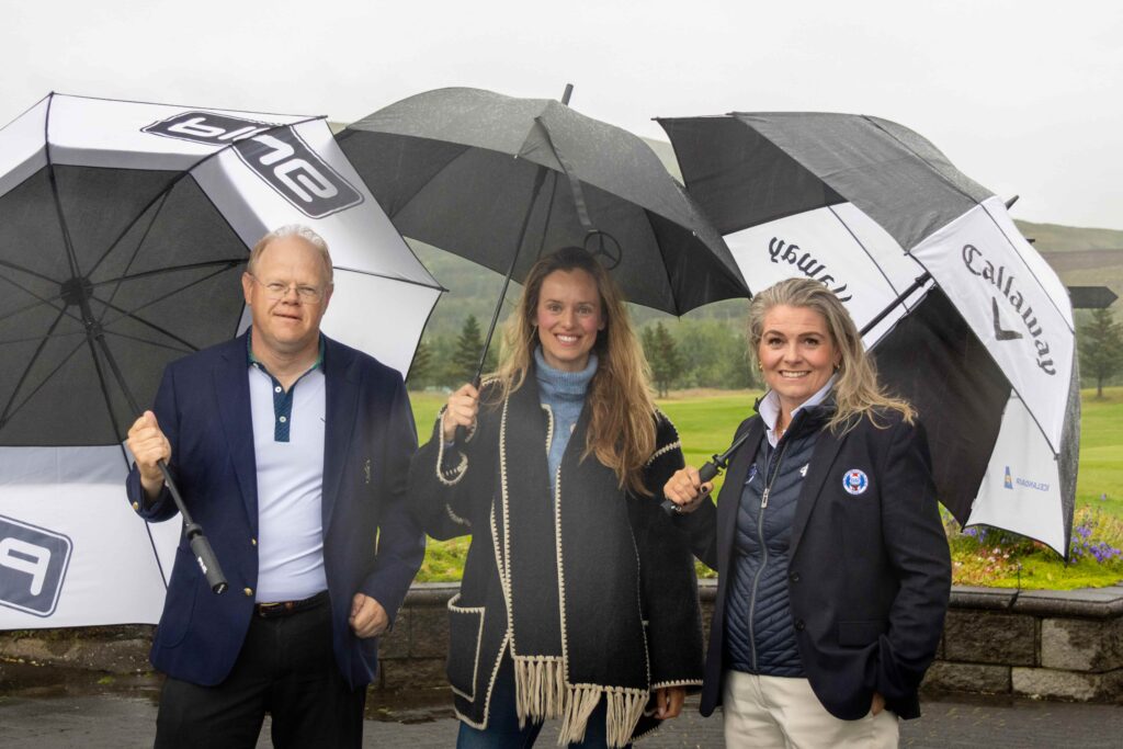 1-499A5338 - Golfsamband Íslands Three adults stand outdoors in the rain, smiling, while holding large black and white umbrellas with logos; grassy field behind them.