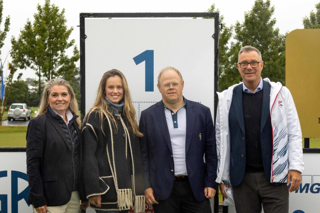 10-499A5435 - Golfsamband Íslands Four adults stand together outdoors in front of a large panel with a blue '1'.