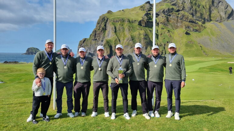 Group of golfers in matching green sweaters and white caps with medals, plus a boy, posing with a trophy on a coastal golf course.