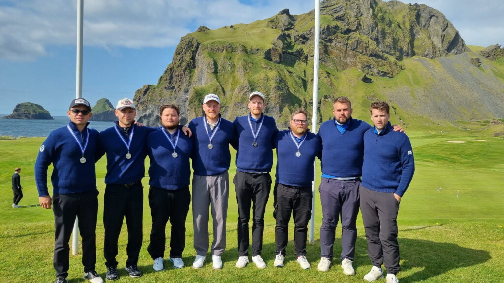 1000017640 1 - Golfsamband Íslands Eight men in matching blue sweaters with silver medals around their necks pose together on a grassy golf course, with a rocky green hillside in the background under a blue sky.