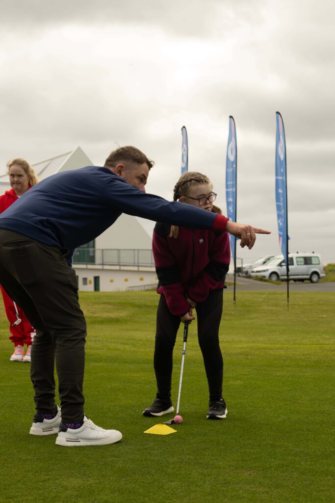 2-499A4738 - Golfsamband Íslands Instructor guides a girl with a cane during a golf drill on a grassy field, with a pink ball on a tee and a yellow marker nearby.