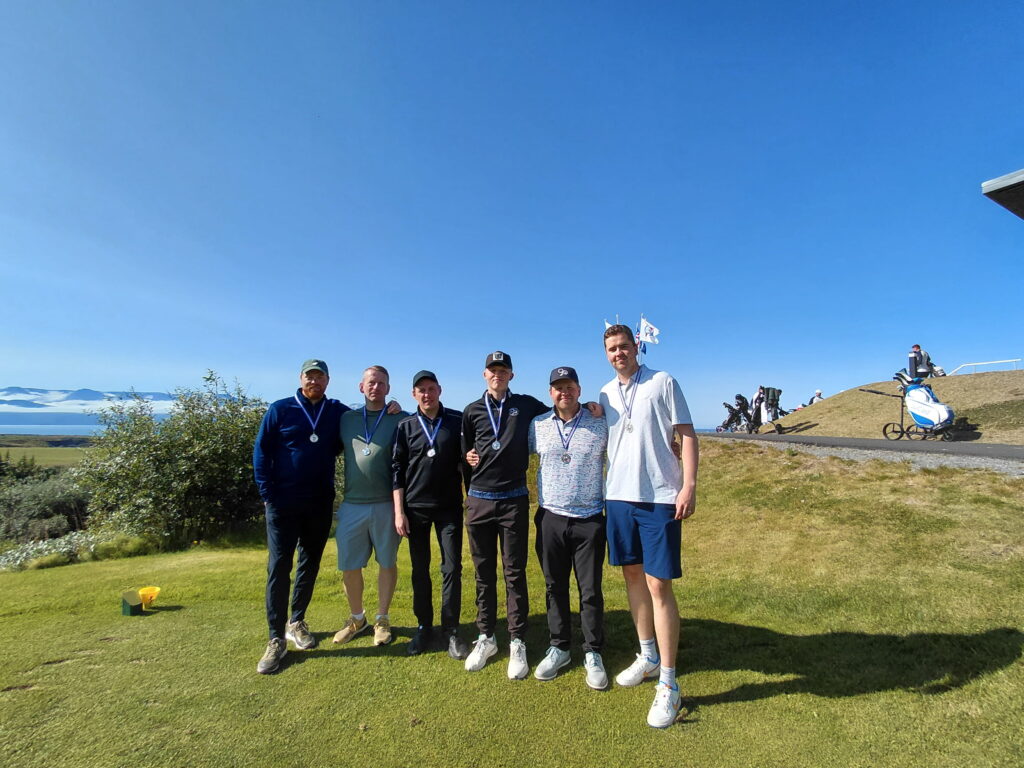 2143 1 - Golfsamband Íslands Seven men stand arm-in-arm on a sunny golf course, each wearing medals after a competition, with hills and equipment visible behind them.