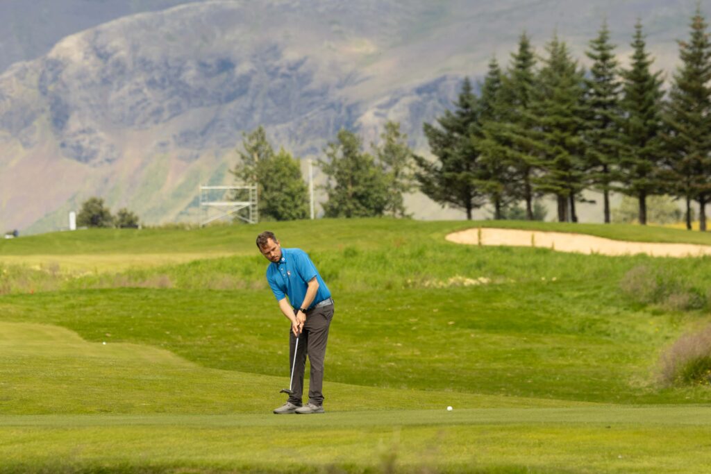 3-499A4810 - Golfsamband Íslands Golfer in a blue shirt preparing to putt on a green golf course with mountains and evergreen trees in the background.