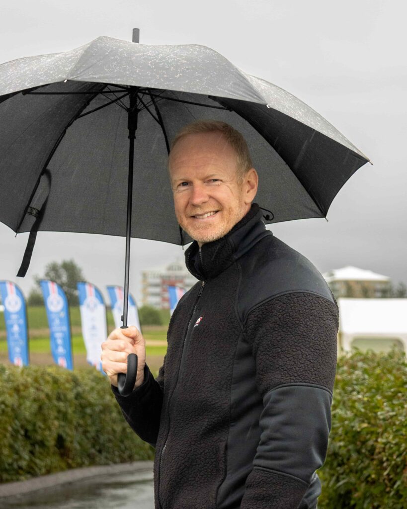 3-499A5352 - Golfsamband Íslands Smiling man in a black jacket holding a large black umbrella in light rain outdoors.