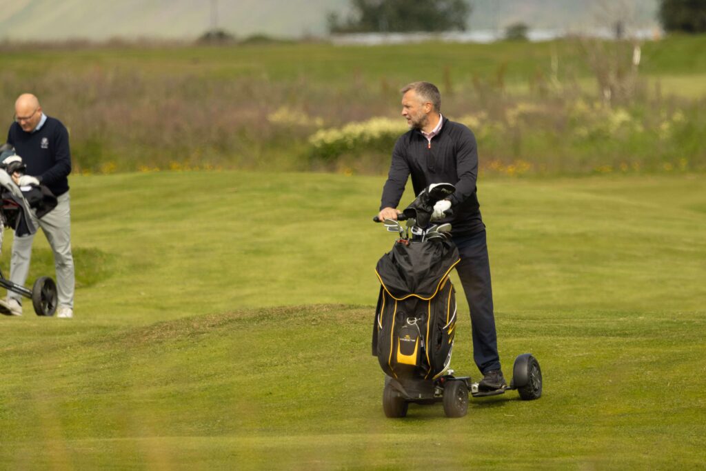 4-499A4819 - Golfsamband Íslands Man in a dark top rides a motorized golf trolley with a large black and yellow bag on a green golf course.
