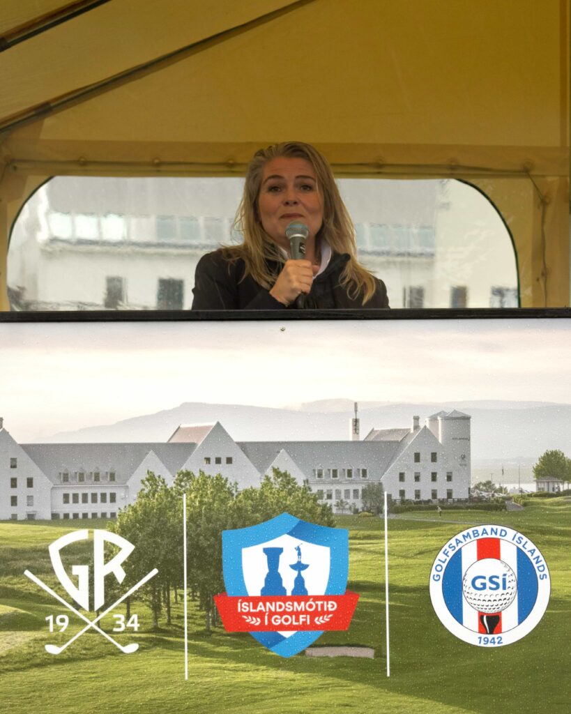 4-499A5363 - Golfsamband Íslands Woman speaks into a microphone from a tent window, with event logos and banners in the foreground.