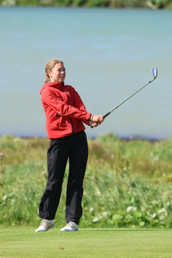 Woman in a red jacket swinging a golf club on a grassy course with a lake in the background.