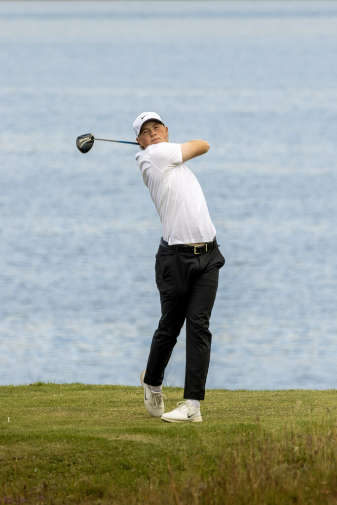 499A4667 1 - Golfsamband Íslands Male golfer in a white polo and cap mid-swing with a driver against a blue lake background.