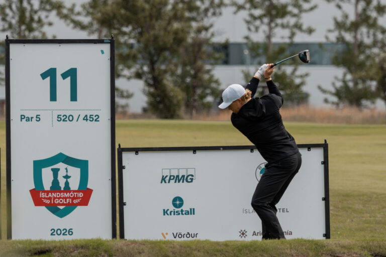 Golfer in a black outfit and white cap mid-swing beside tournament signs on a golf course.