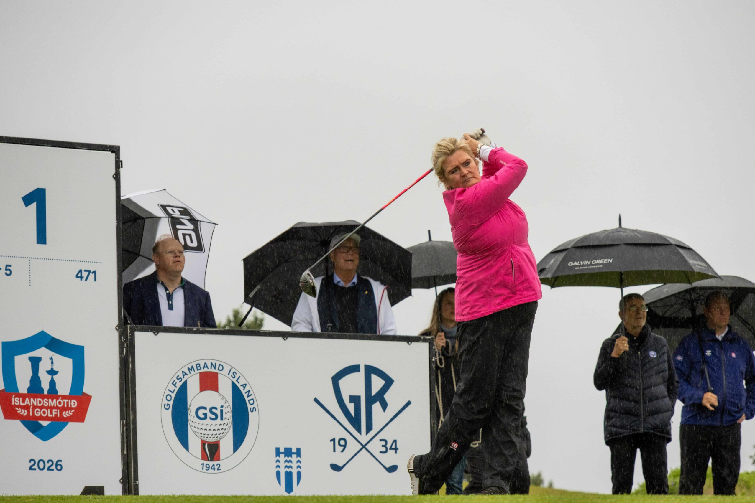 Female golfer in bright pink jacket swings a club while spectators hold umbrellas in the rain.