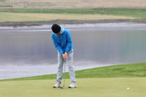 Golfer in a blue jacket and black beanie lining up a putt on a green, with water and landscape in the background.