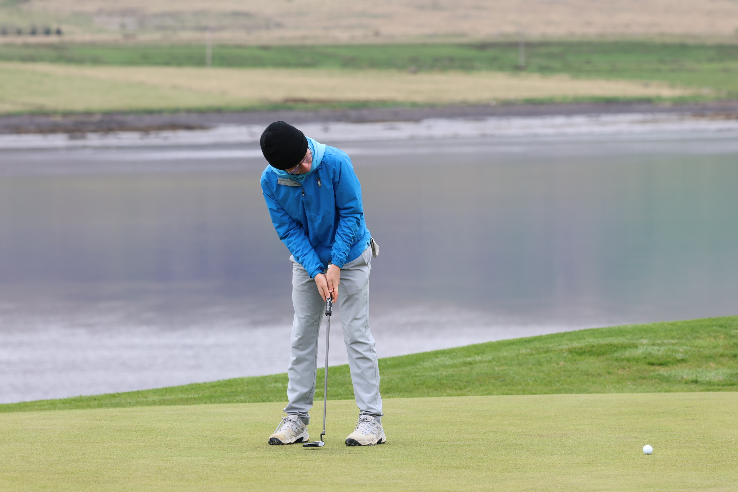 Golfer in a blue jacket and black beanie lining up a putt on a green, with water and landscape in the background.