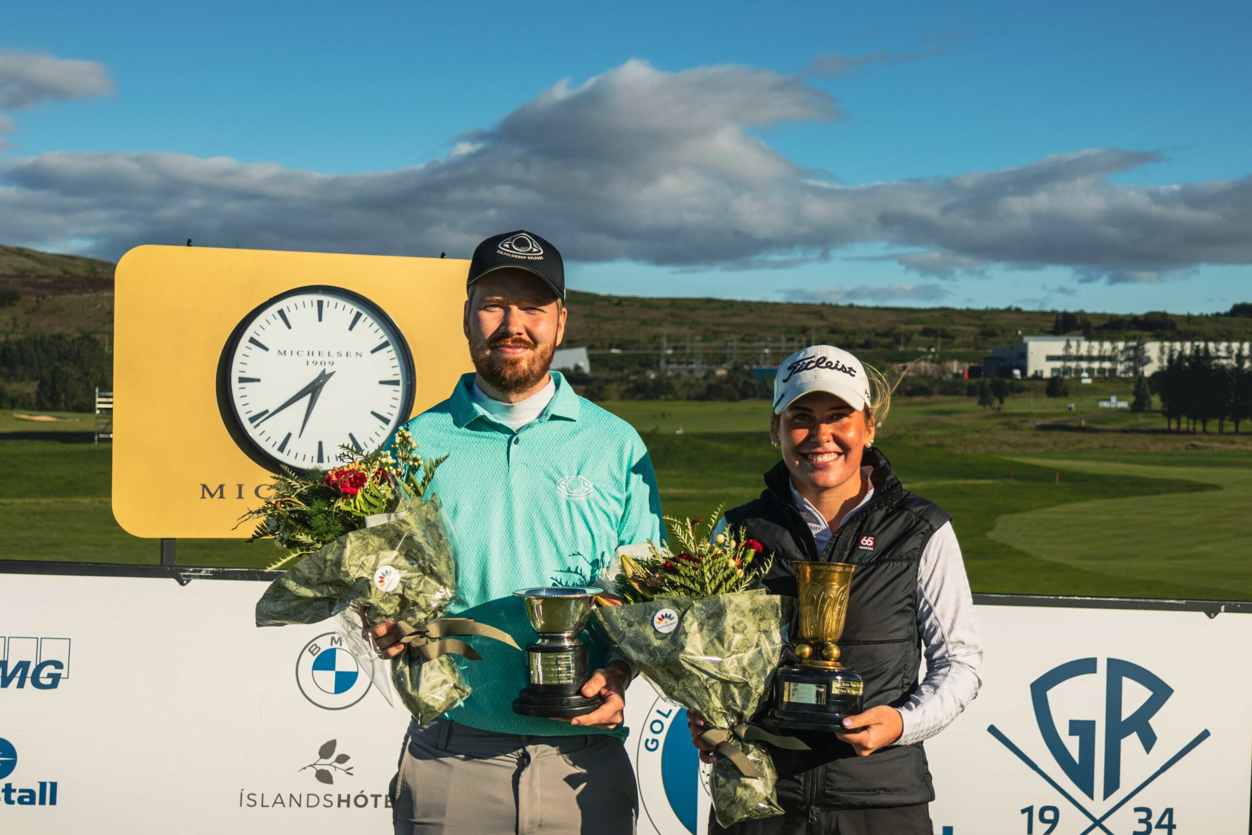 Two golfers on a podium posing with trophies and bouquets outdoors, in front of a large yellow clock sign under a blue sky.