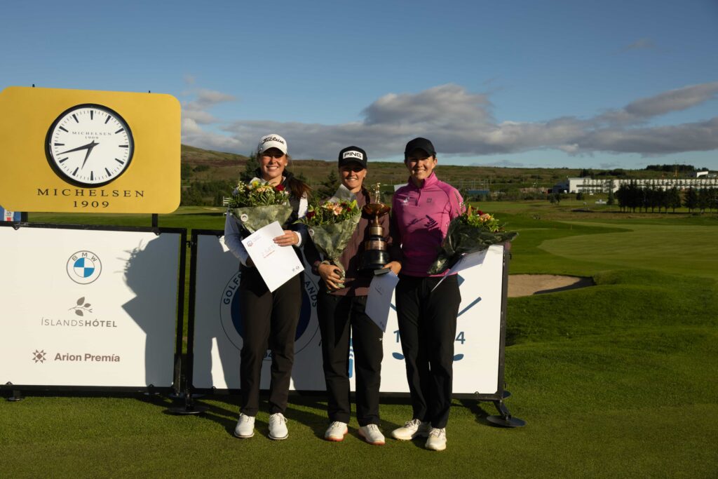 499A9108 1 - Golfsamband Íslands Three female golfers on a podium smiling, holding flowers and a trophy, with a yellow sign and clock in the background at a sunny golf course.