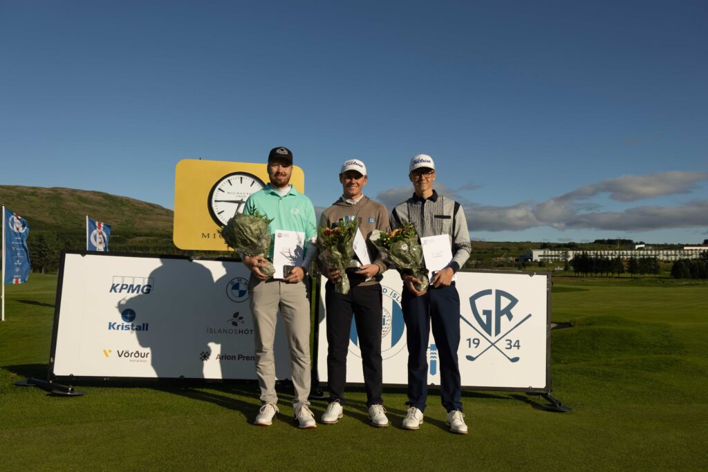 499A9135 1 - Golfsamband Íslands Three men on a winners' podium pose with bouquets and certificates at an outdoor golf event, blue sky above and sponsor signs behind them.