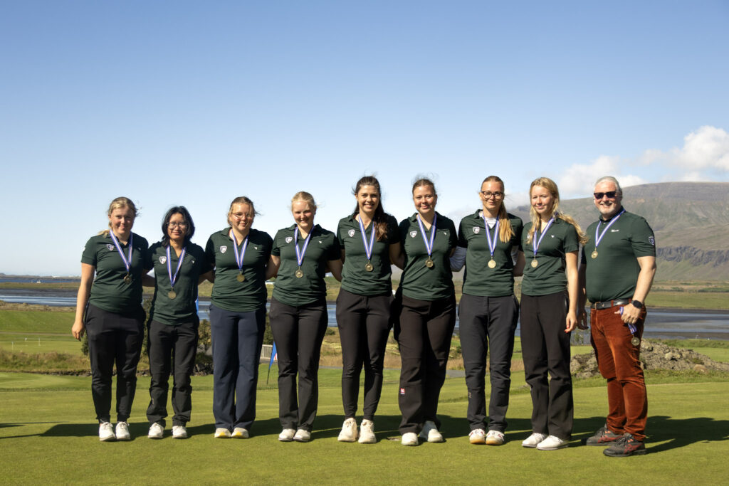 499A9566 - Golfsamband Íslands Group of athletes in matching green shirts with medals posing for a team photo outdoors against a blue sky and hills in the background.