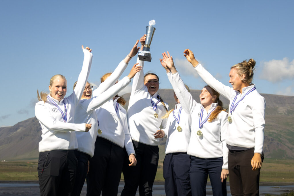 499A9701 - Golfsamband Íslands Group of young women in white jackets lift a trophy together, celebrating victory outdoors with medals around their necks.
