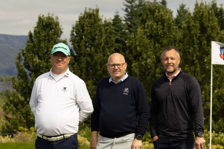 Three men stand together outdoors on a golf course, posing for a photo with trees and a flag in the background.