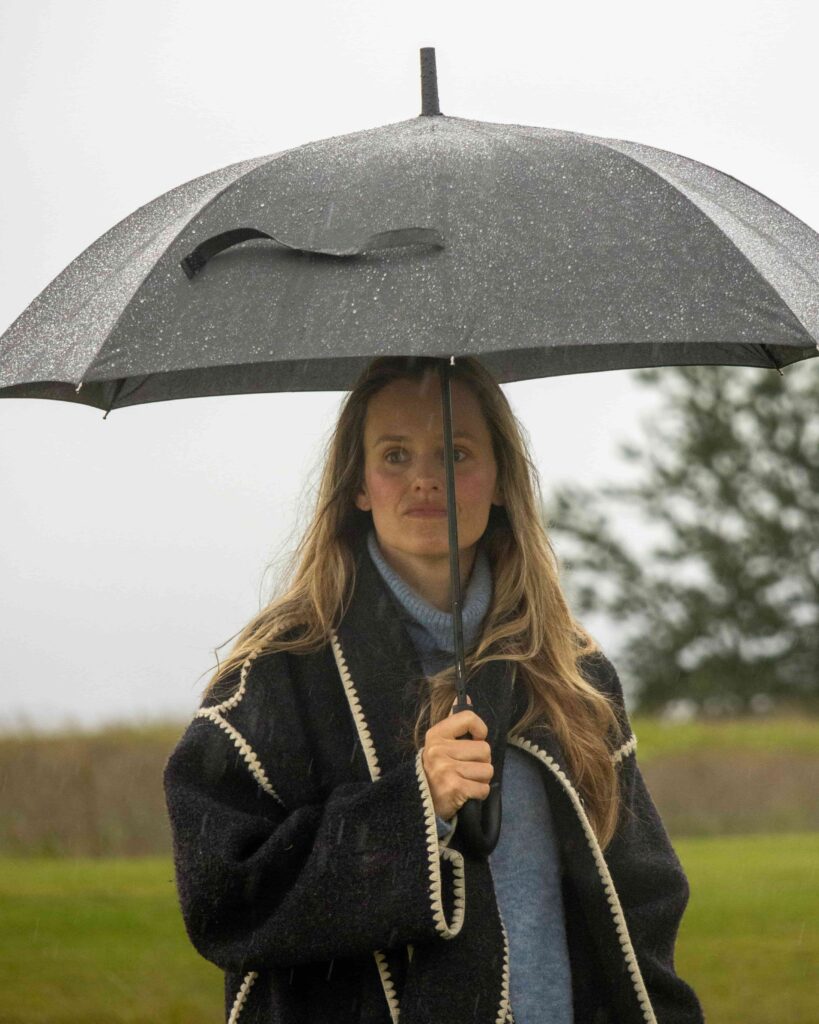 5-499A5372 - Golfsamband Íslands Woman standing outdoors in the rain, holding a black umbrella with droplets on its surface.