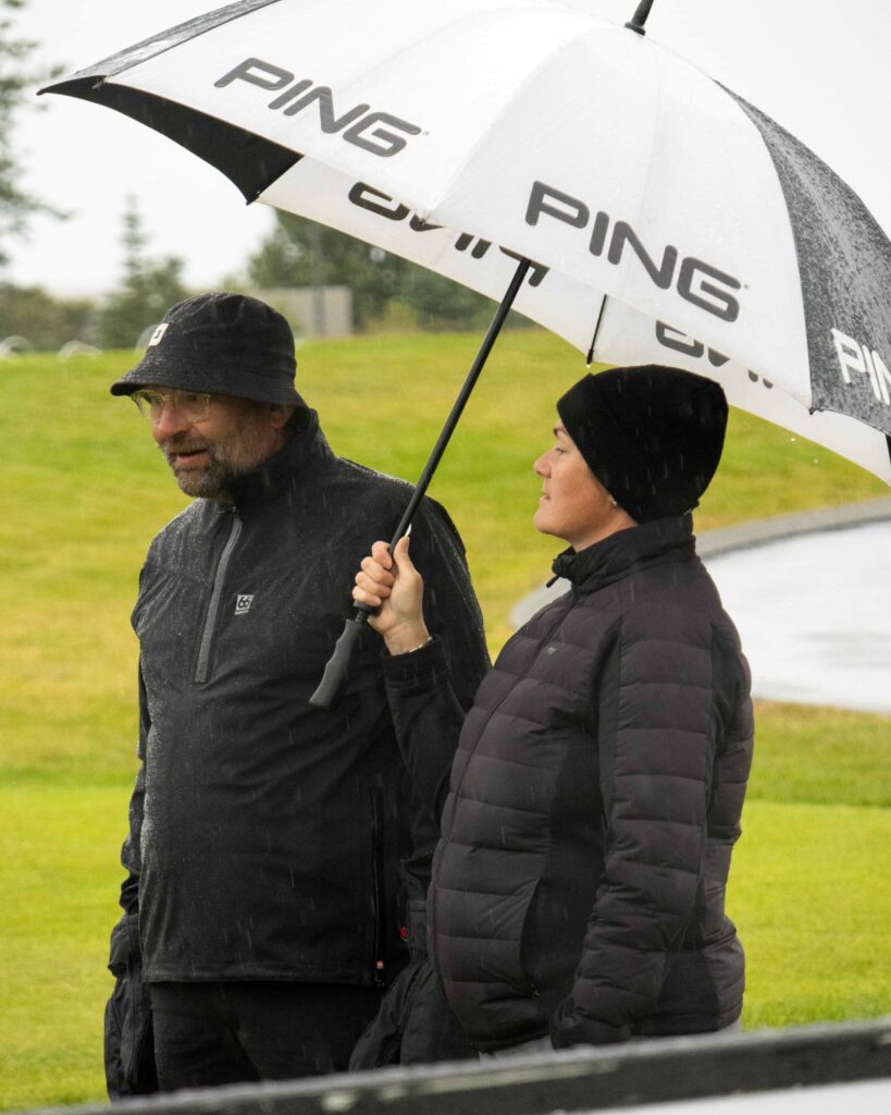 6-499A5378 - Golfsamband Íslands Two people standing in a rainy outdoor field, sharing a white umbrella with the PING logo; man in a black rain jacket and hat, woman in a black puffer jacket.