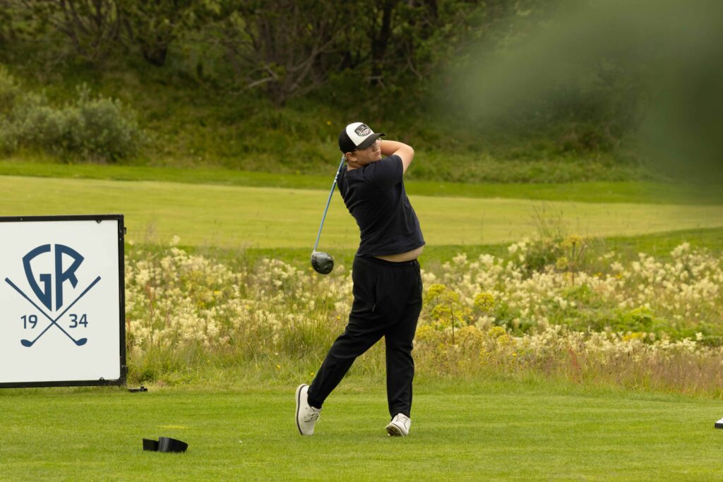 7-499A5161 - Golfsamband Íslands Golfer mid-swing on a grassy golf course, wearing a dark shirt and cap, with a sign featuring a crest on the left.
