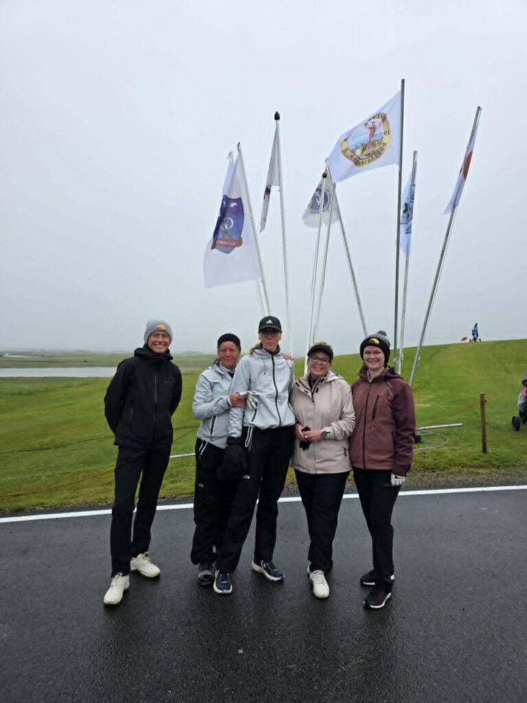 777800997_1451687376768196_1567842844244177885_n - Golfsamband Íslands Five people in jackets posing on a wet road with tall flag poles and flags fluttering in the background.