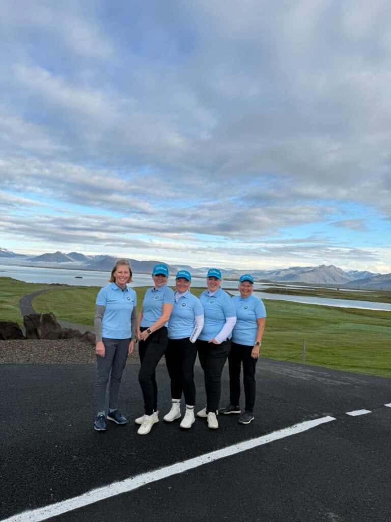 777804769_1451687606768173_8430285830680971311_n - Golfsamband Íslands Five women in matching light-blue polo shirts and caps pose outdoors with a mountainous, lakeside landscape in the background.