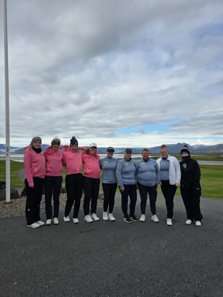 777854205_1451687026768231_2912726325192613290_n - Golfsamband Íslands Group of people in pink and blue jackets posing outdoors on a paved area with a lake and mountains in the background, under a cloudy sky.