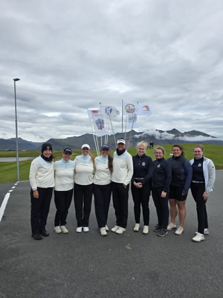 777868144_1451687140101553_4880898309684812951_n - Golfsamband Íslands Group of athletes posing together on a road with flags and mountains in the background under a cloudy sky, all wearing athletic jackets and pants.
