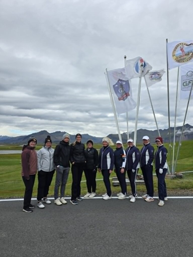 777881635_1451687223434878_8453027248128250538_n - Golfsamband Íslands Group of athletes standing in a line outdoors, with several tall flags and a cloudy sky behind them, hills in the distance.