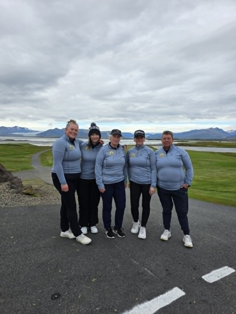 778401384_1451687103434890_2130161585196580766_n - Golfsamband Íslands Five people in matching light-blue jackets pose together on a paved road with green fields and distant mountains under a cloudy sky.