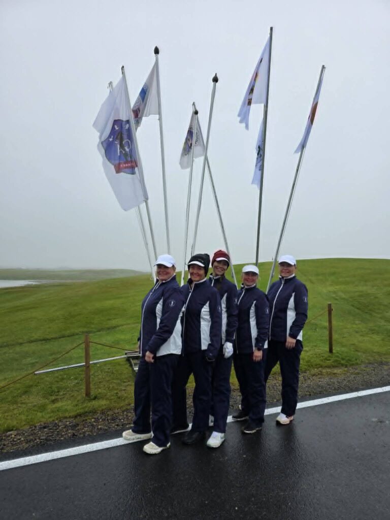 778565335_1451687560101511_1942323163740991544_n - Golfsamband Íslands Group of seven people in matching navy jackets and white caps carrying tall flagpoles with white flags outdoors on a grassy area by the road.