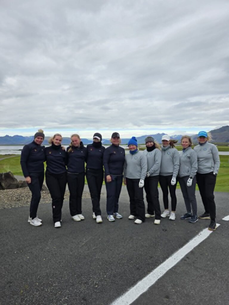 779756291_1451687090101558_4651613719176102733_n - Golfsamband Íslands Ten people in athletic jackets standing shoulder to shoulder on a paved road outdoors, with a cloudy sky and hills in the background.