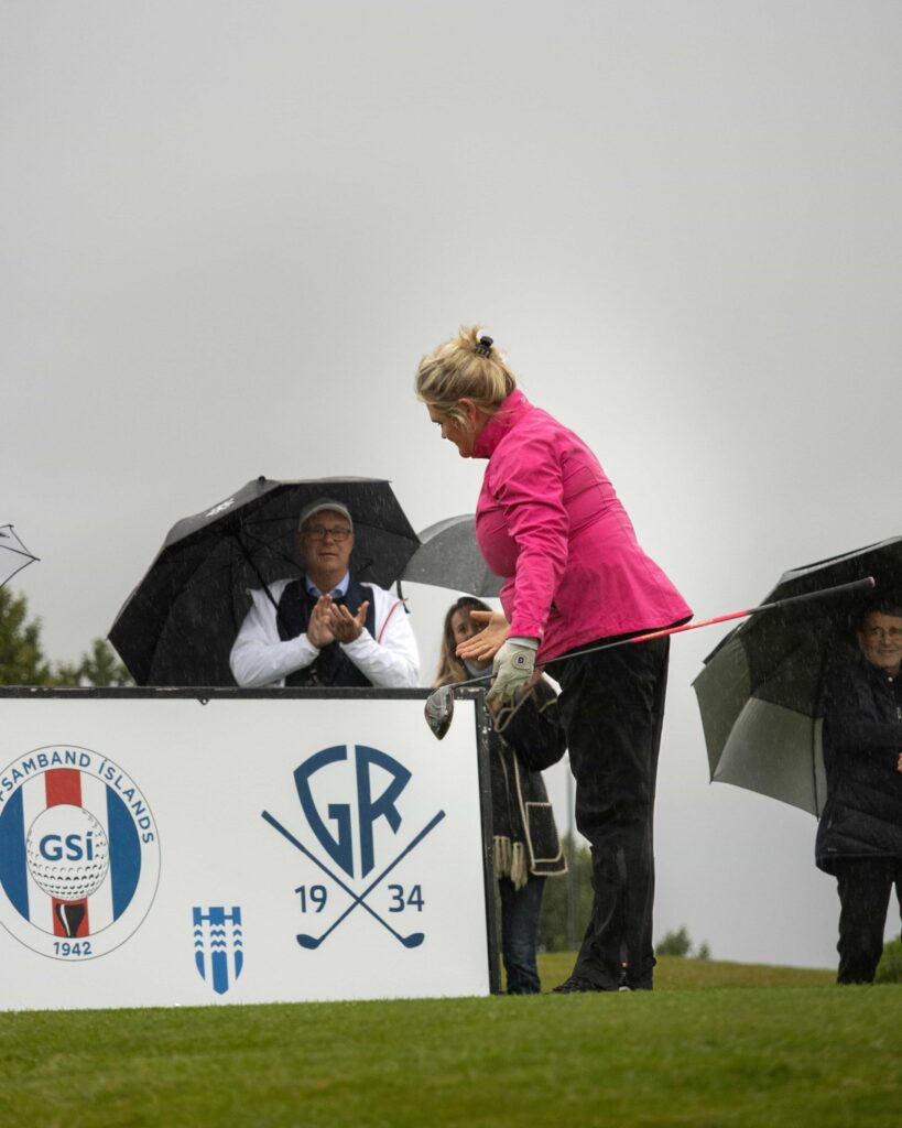 8-499A5401 - Golfsamband Íslands Woman in a bright pink jacket leaning over a podium during a rainy golf event, spectators with umbrellas applaud nearby; sponsor logos visible on the barrier.