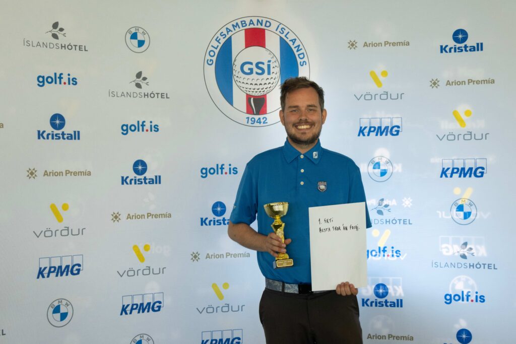 9-499A5212 - Golfsamband Íslands Award recipient in a blue polo shirt holding a trophy and certificate in front of a sponsor backdrop.