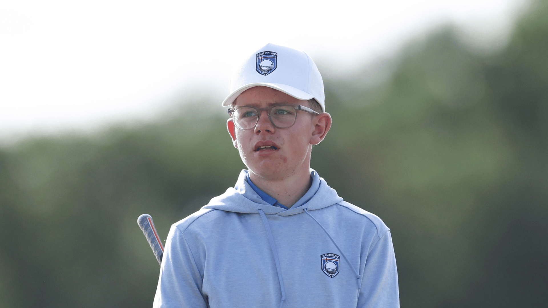 Young person wearing a white cap with a Team Iceland logo and a light blue hoodie, holding a hockey stick outdoors.