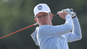 Golfer wearing a white cap with a blue emblem and light blue jacket, in the middle of a swing.