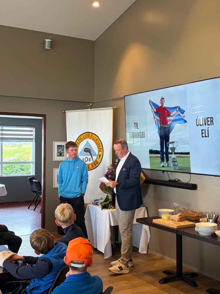 IMG_8642 - Golfsamband Íslands Speaker in a navy blazer presenting to a seated group of young athletes, banners and a large screen behind him.