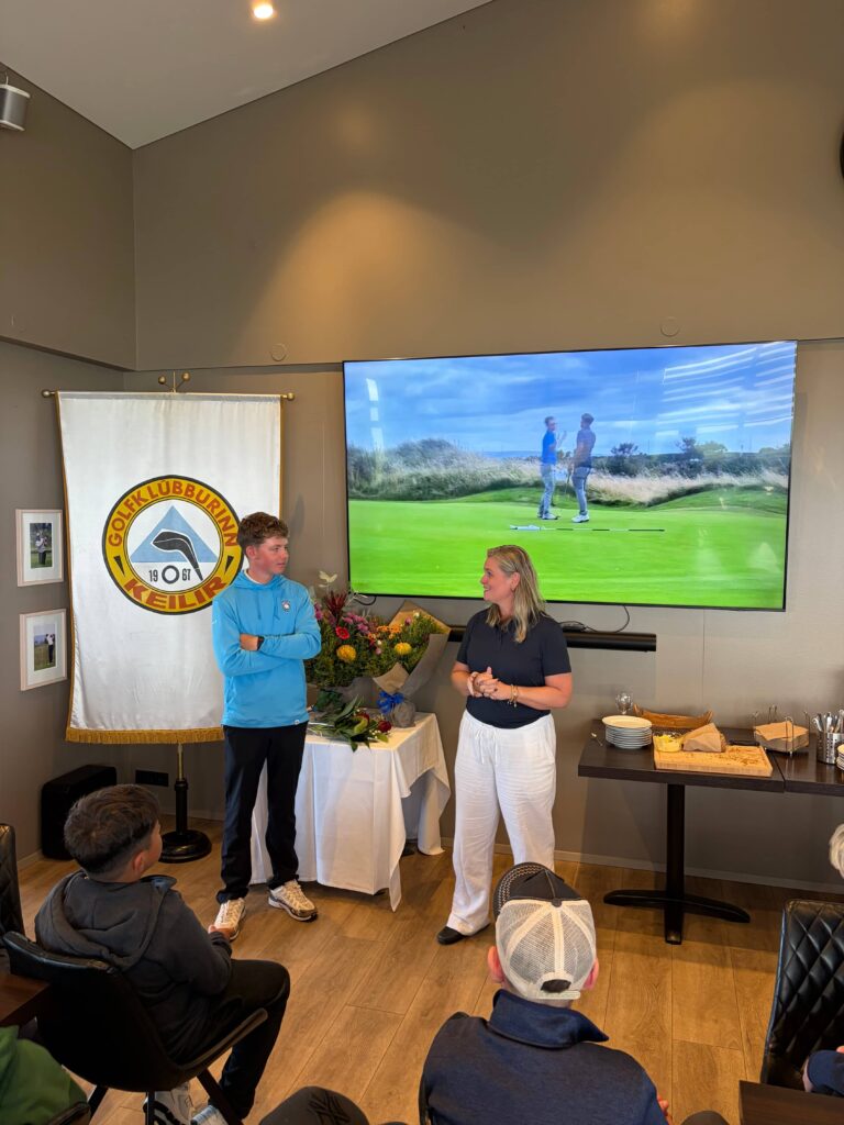 IMG_8644 - Golfsamband Íslands Two people speak to a small audience in a room with a large golf-themed banner and a TV screen showing a golf course.