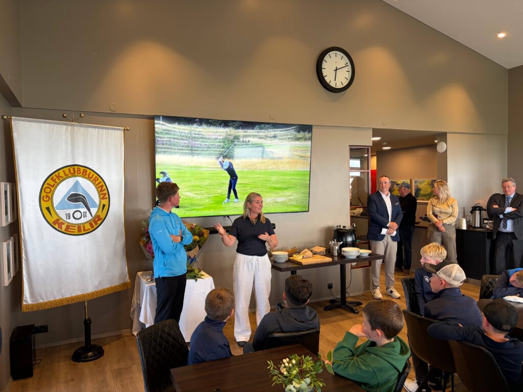 IMG_8646 - Golfsamband Íslands Group of people at a club event: presenters speak near a large TV showing a golfer on a green, with a banner on the left and a clock on the wall behind.